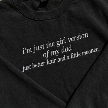 Close-up of a black crewneck t-shirt featuring white embroidered text that reads 'i'm just the girl version of my dad just better hair and a little meaner.'.