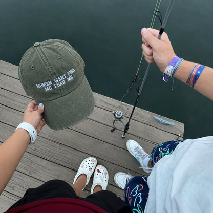 An olive green baseball cap with white embroidered text reading 'WOMEN WANT FISH ME FEAR ME' is held up by a person on a wooden dock over dark water, with a fishing rod and the person's legs and hands visible.