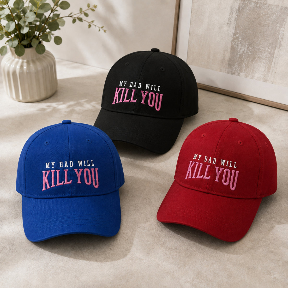 Three baseball caps displayed on a light beige surface: one royal blue, one black, and one red. Each cap has embroidered text reading 'MY DAD WILL KILL YOU' in contrasting white and pink lettering.