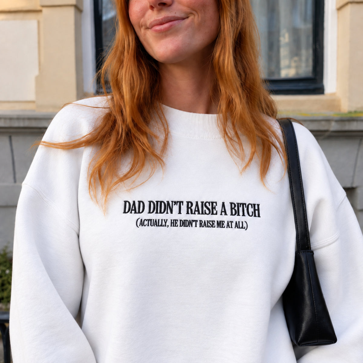A woman wearing a white crewneck sweatshirt with black printed text that reads 'DAD DIDN'T RAISE A BITCH (ACTUALLY, HE DIDN'T RAISE ME AT ALL)'. She is outdoors in front of a building, carrying a black shoulder bag.
