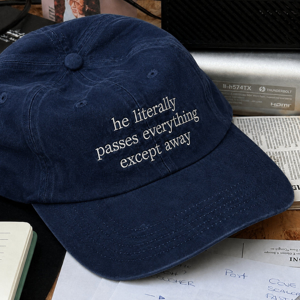 A navy blue dad hat with white embroidered text reading 'he literally passes everything except away', shown resting on a desk with papers and electronic equipment in the background.