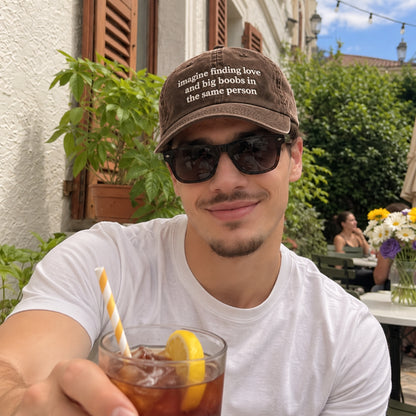 A man wearing a brown distressed baseball cap with the text 'Imagine finding love and big boobs in the same person' and black sunglasses, sitting at an outdoor cafe and holding an iced drink with a lemon slice.