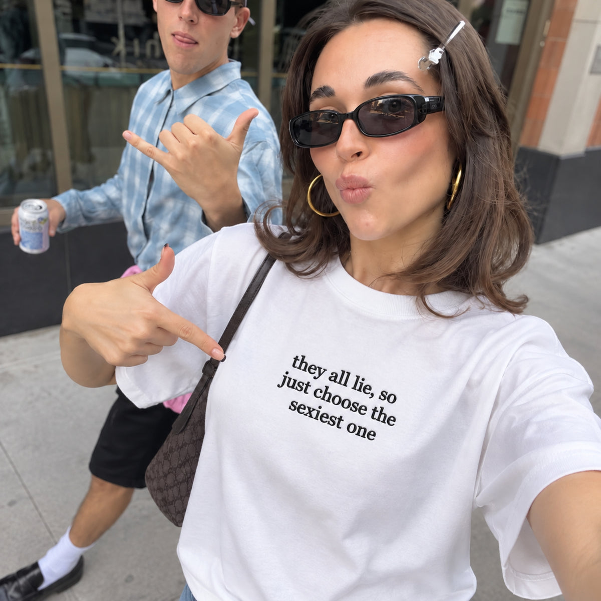 Woman wearing a white embroidered T-shirt featuring the funny quote “they all lie, so just choose the sexiest one” in black stitching, posing for a street-style selfie with sunglasses and pointing at the chest embroidery.