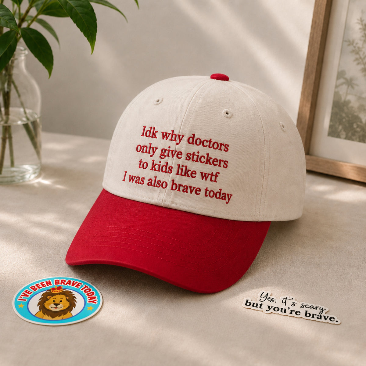 A baseball cap with a white crown and red brim, featuring red embroidered text that reads 'Idk why doctors only give stickers to kids like wtf I was also brave today'. A matching round sticker and a small script sticker are placed beside it on a light surface with soft shadows, alongside a plant and framed picture in the background.