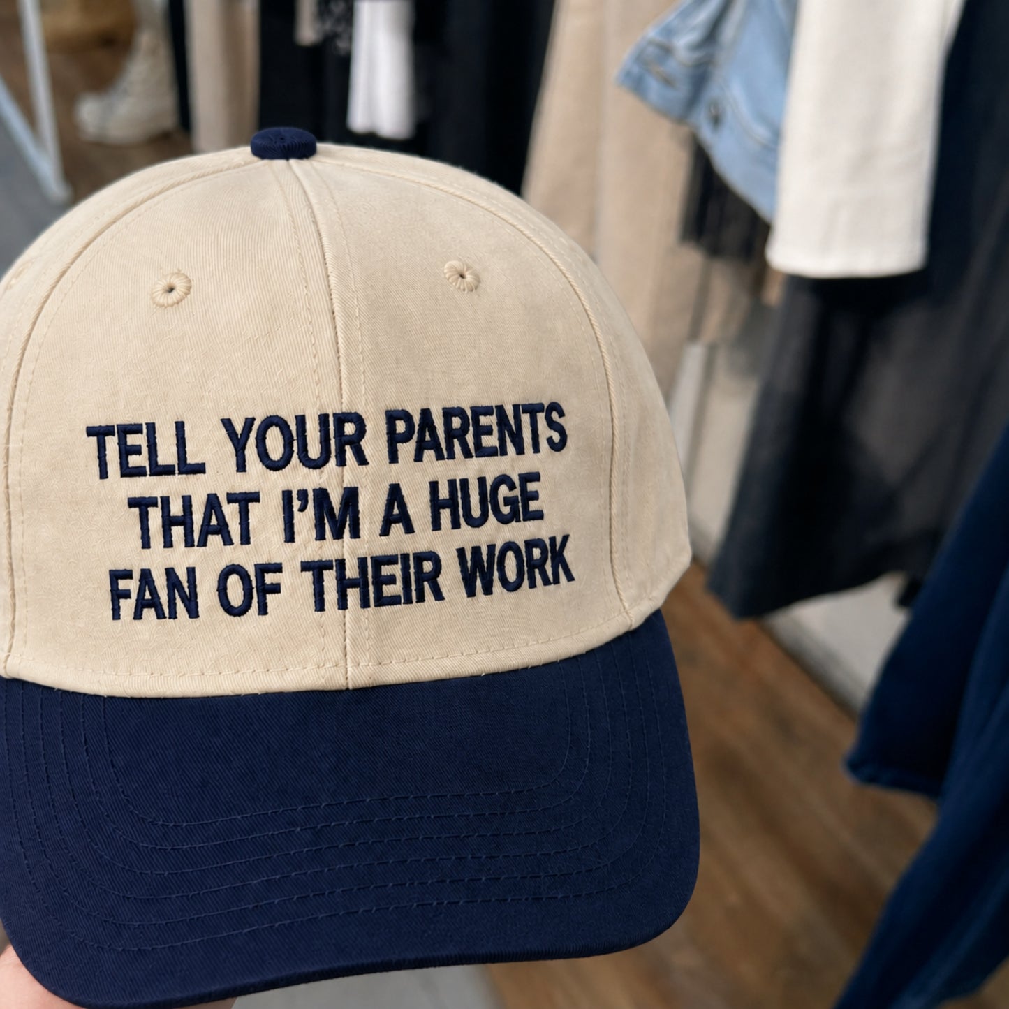 A beige and navy blue baseball cap held up in a store setting, featuring the embroidered text 'TELL YOUR PARENTS THAT I'M A HUGA FAN OF THEIR WORK' on the front panel.