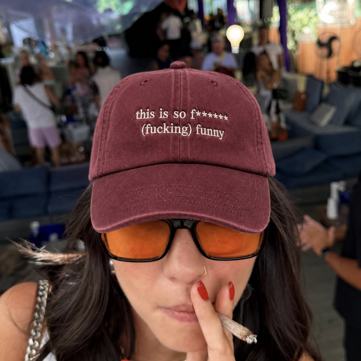 A close-up lifestyle shot of a woman wearing a maroon baseball cap with white embroidered text that reads 'this is so f***** (fucking) funny'. She is also wearing orange-tinted sunglasses and holding a cigarette in a crowded, outdoor event setting.