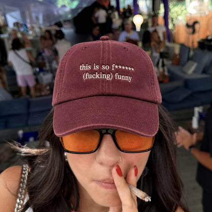 A close-up lifestyle shot of a woman wearing a maroon baseball cap with white embroidered text that reads 'this is so f***** (fucking) funny'. She is also wearing orange-tinted sunglasses and holding a cigarette in a crowded, outdoor event setting.