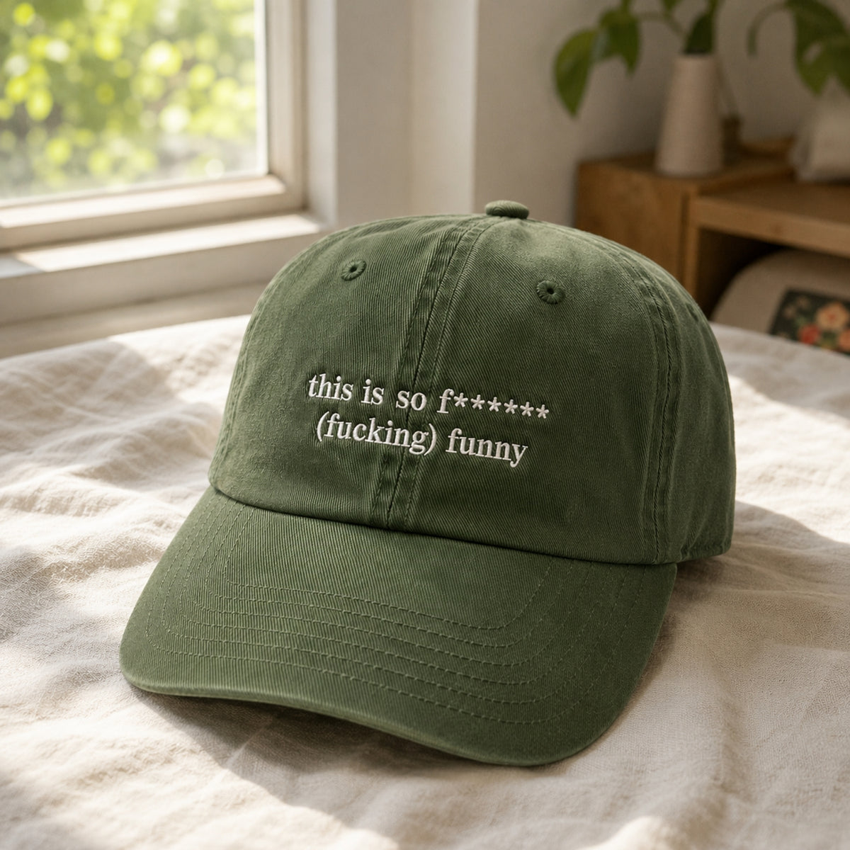 An olive green dad hat baseball cap with white embroidered text reading 'this is so f****** funny' resting on a white textured bedspread in a softly lit bedroom.