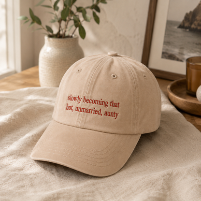 A beige baseball cap with the phrase 'slowly becoming that hot, unmarried, aunty' embroidered in red on the front, resting on a textured beige fabric in a warm, styled interior setting.