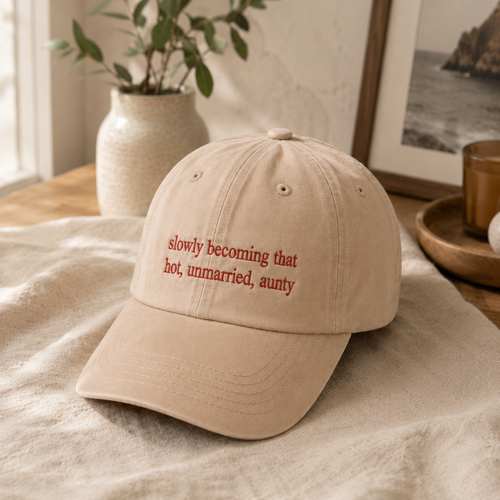 A beige baseball cap with the phrase 'slowly becoming that hot, unmarried, aunty' embroidered in red on the front, resting on a textured beige fabric in a warm, styled interior setting.