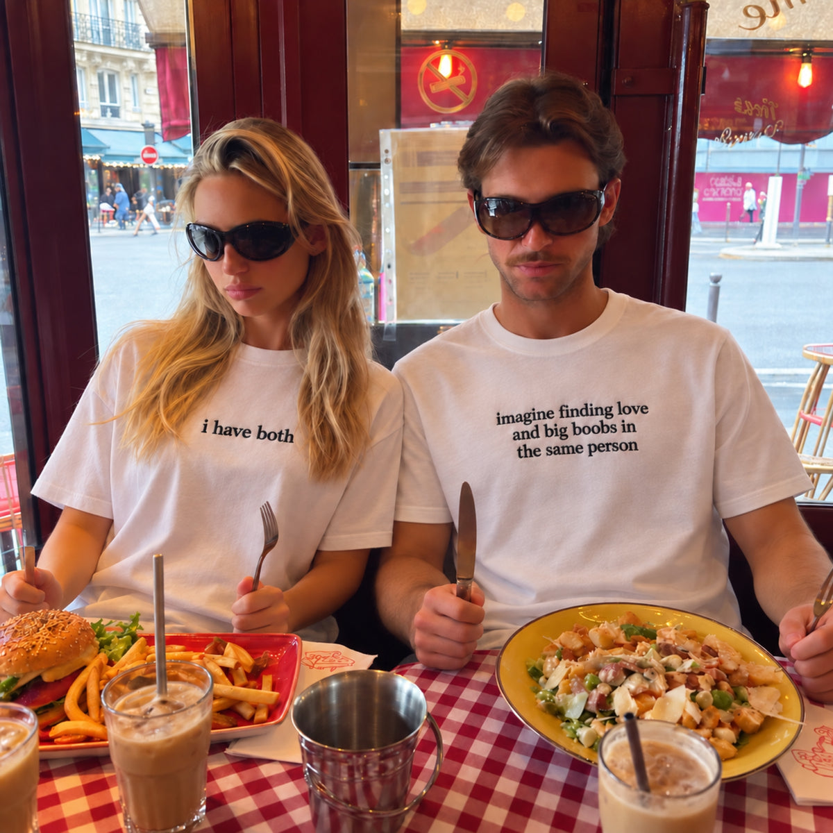 A man and a woman sitting at a restaurant table wearing matching white graphic t-shirts with black text. The woman's shirt reads 'i have both' and the man's shirt reads 'imagine finding love and big boobs in the same person'.