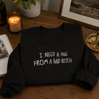 A black crewneck sweatshirt with the text 'I NEED A HUG FROM A BAD BITCH' printed in white across the chest, displayed on a wooden table surrounded by cozy lifestyle props like a lit candle, framed picture, and plant.