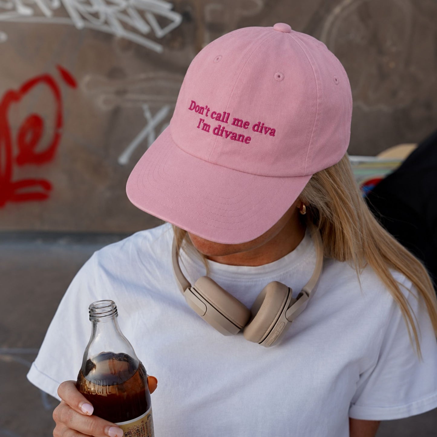 A person wearing a light pink baseball cap with the embroidered text 'Don't call me diva I'm divane' in darker pink. The person is also wearing a white t-shirt and beige over-ear headphones around their neck, while holding a glass bottle of soda.