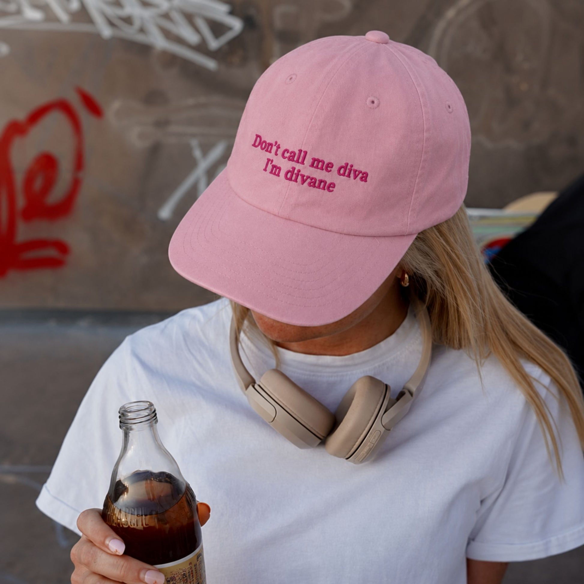 A person wearing a light pink baseball cap with the embroidered text 'Don't call me diva I'm divane' in darker pink. The person is also wearing a white t-shirt and beige over-ear headphones around their neck, while holding a glass bottle of soda.