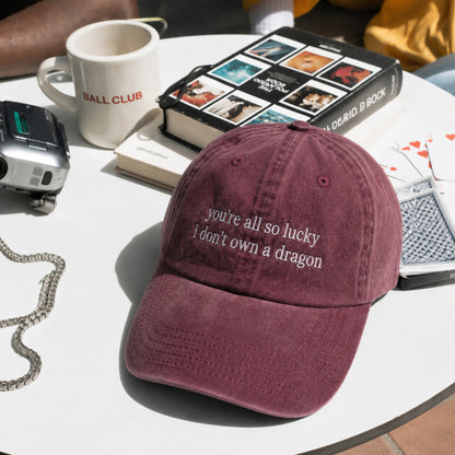 A dusty burgundy baseball cap with white embroidered text reading 'you're all so lucky I don't own a dragon' sits on a white table surrounded by lifestyle props including a 'BALL CLUB' mug, a camera, a book, and playing cards.