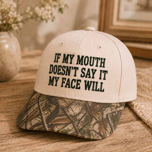 A beige baseball cap with black embroidered text reading 'IF MY MOUTH DOESN'T SAY IT MY FACE WILL' and a camouflage-patterned brim, displayed on a wooden surface with a blurred indoor background.