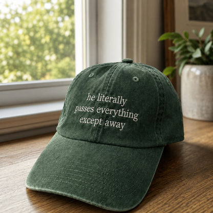 A dark green washed baseball cap with white embroidered text reading 'he literally passes everything except away', resting on a wooden table near a window with a potted plant in the background.