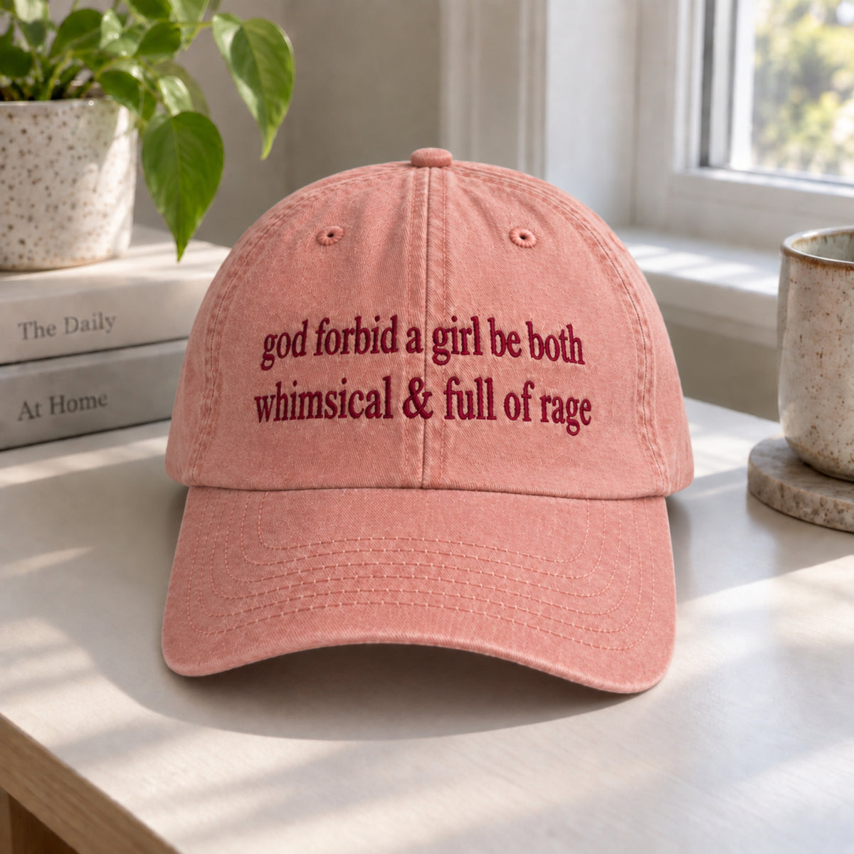 A dusty pink baseball cap with dark red embroidered text reading 'god forbid a girl be both whimsical & full of rage', displayed on a light wooden table in a bright room with a plant, books, and a mug in the background.