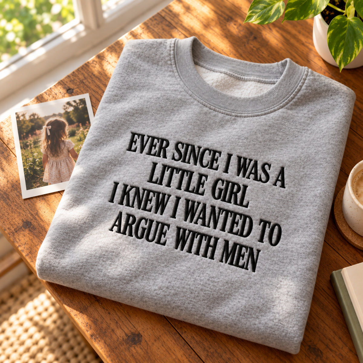 A folded light gray crewneck sweatshirt with black text reading 'EVER SINCE I WAS A LITTLE GIRL I KNEW I WANTED TO ARGUE WITH MEN' lies on a wooden table next to a photo print and a mug.
