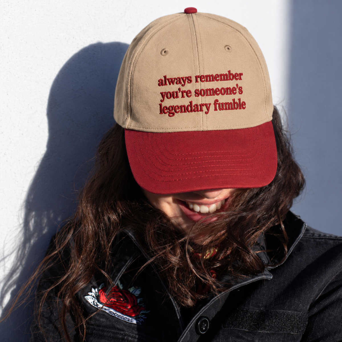 A person wearing a beige baseball cap with a red brim and red embroidered text that reads 'always remember you're someone's legendary fumble'. The person is smiling, with long brown hair blowing in the wind, against a white wall with a strong shadow.