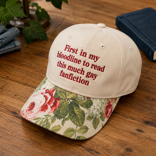 A baseball cap placed on a wooden table, with a cream crown and a floral brim featuring red roses and green leaves. The cap has red embroidered text reading, 'First in my bloodline to read this much gay fanfiction.