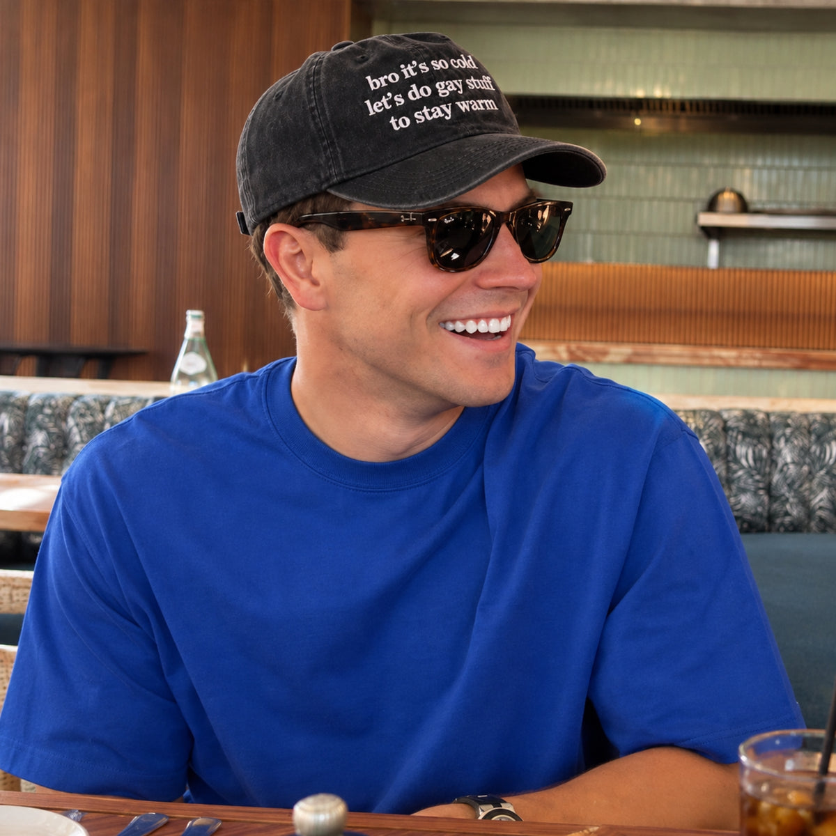 A man wearing a black baseball cap with white embroidered text, dark sunglasses, and a bright blue t-shirt, sitting in a restaurant setting.