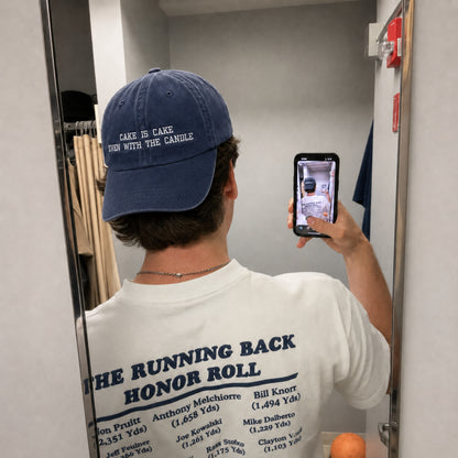 A person taking a mirror selfie in a fitting room, wearing a navy blue baseball cap with white embroidered text that reads 'CAKE IS CAKE EAT WITH THE CANDLE' and a white graphic T-shirt.