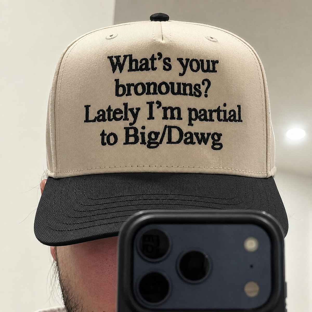 A beige and black trucker-style cap with embroidered text reading 'What's your bronouns? Lately I'm partial to Big/Dawg', shown in a mirror selfie with a phone partially covering the bottom.