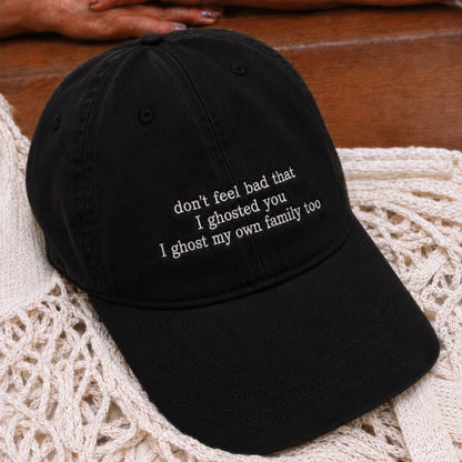 A black baseball cap with white embroidered text reading 'don't feel bad that I ghosted you I ghost my own family too', resting on a cream crochet blanket over a wooden surface.