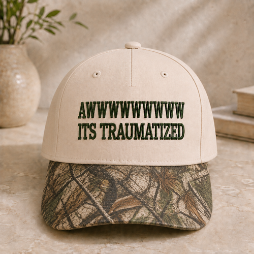 A beige baseball cap with dark green embroidered text reading 'AWWWWWWWWW ITS TRAUMATIZED' on the front, featuring a camouflage-patterned brim, displayed on a stone surface with a vase and books in the background.