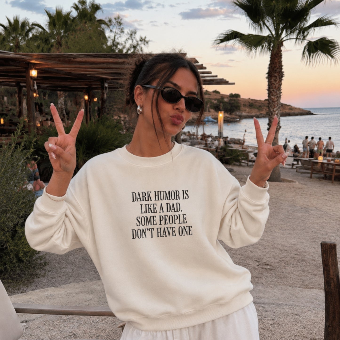A woman posing outdoors at a beachside restaurant during sunset, wearing a cream-colored sweatshirt with black text that reads 'DARK HUMOR IS LIKE A DAD, SOME PEOPLE DON'T HAVE ONE'.