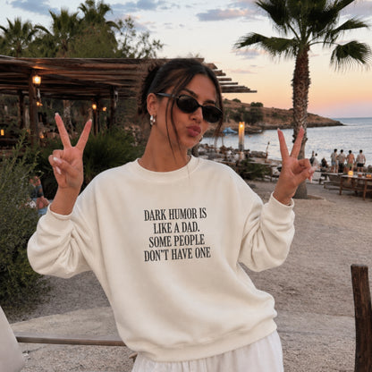 A woman posing outdoors at a beachside restaurant during sunset, wearing a cream-colored sweatshirt with black text that reads 'DARK HUMOR IS LIKE A DAD, SOME PEOPLE DON'T HAVE ONE'.