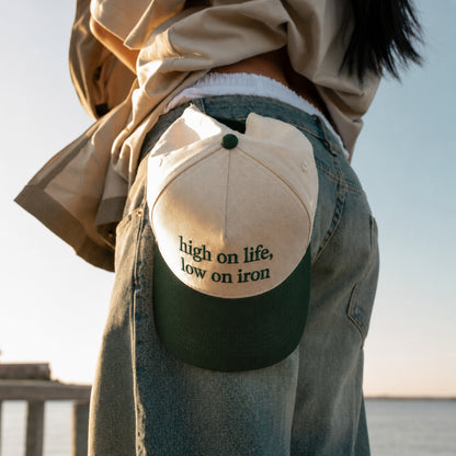 A person wearing a beige and dark green trucker-style cap with the embroidered text 'high on life, low on iron' tucked into the back pocket of their blue jeans.