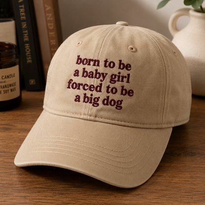 A beige baseball cap with dark red embroidered text reading 'born to be a baby girl forced to be a big dog' resting on a wooden table with books and a ceramic vase in the background.