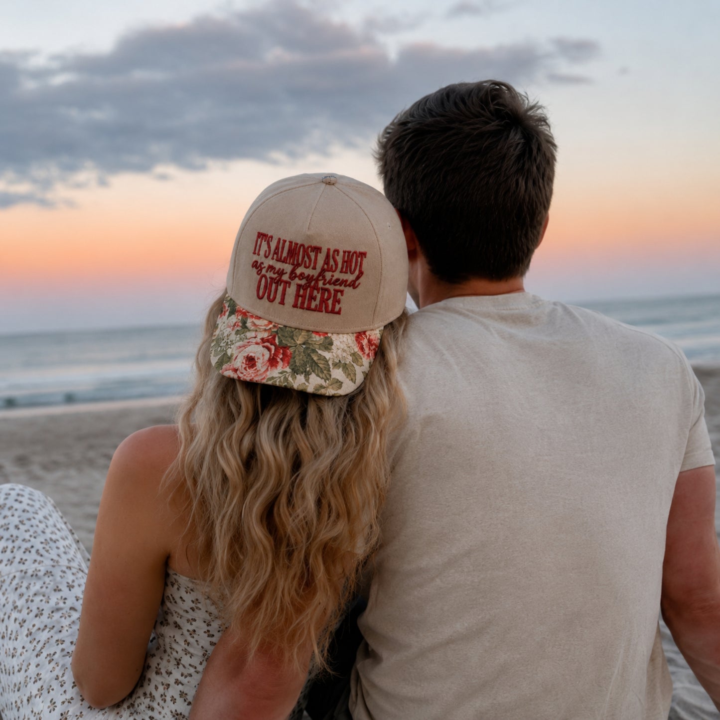 A couple sitting together on a beach at sunset, viewed from behind. The woman wears a beige baseball cap with red embroidered text reading 'IT'S ALMOST AS HOT as my body gets OUT HERE' and a floral brim, while the man wears a light beige T-shirt.