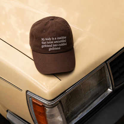 A dark brown baseball cap with white embroidered text reading 'My body is a machine that turns uncuddled, girlfriend into cuddled girlfriend' resting on the front fender of a beige vintage car.