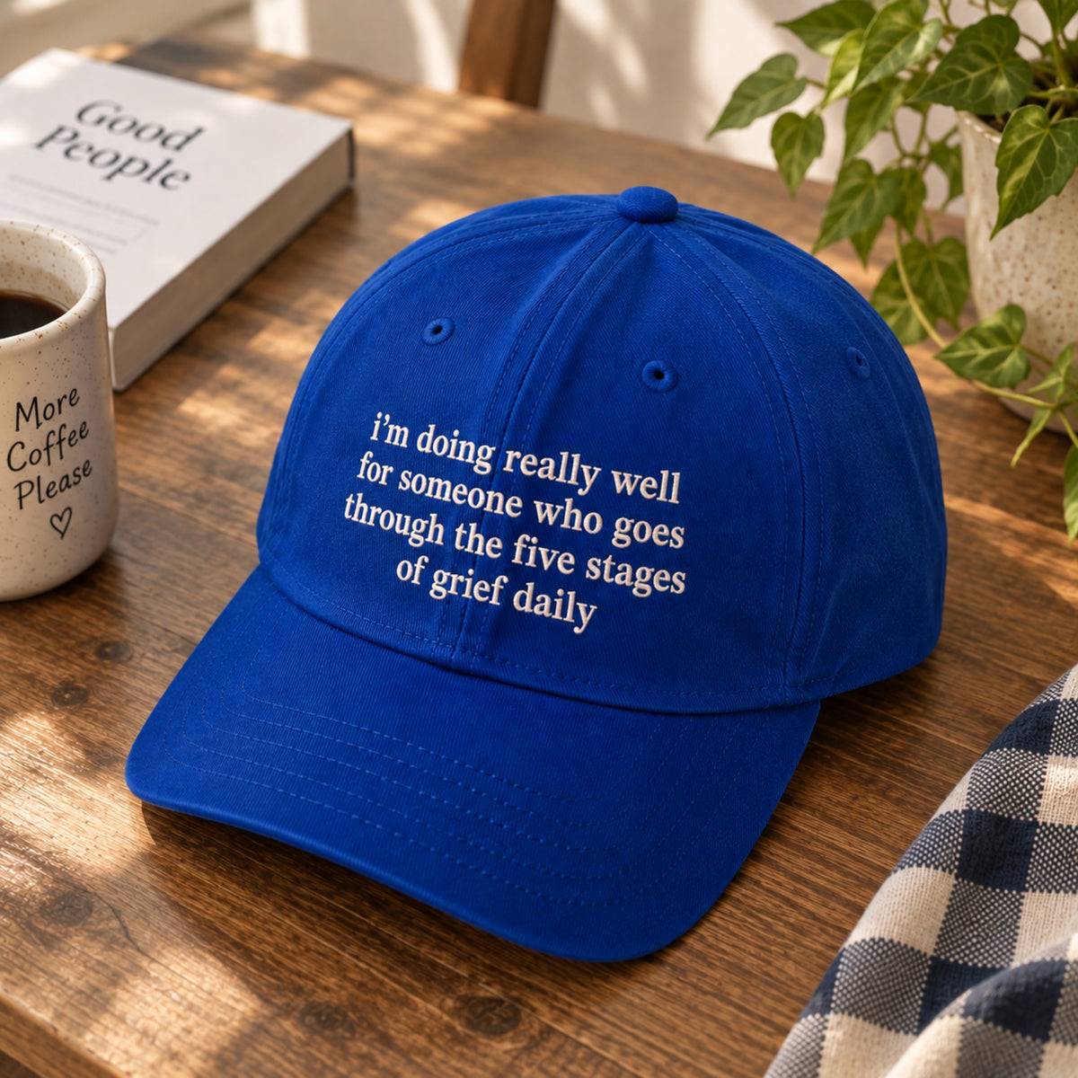 A royal blue baseball cap with white embroidered text reading 'I'm doing really well for someone who goes through the five stages of grief daily', displayed on a wooden table next to a coffee mug, a book, and a plant.