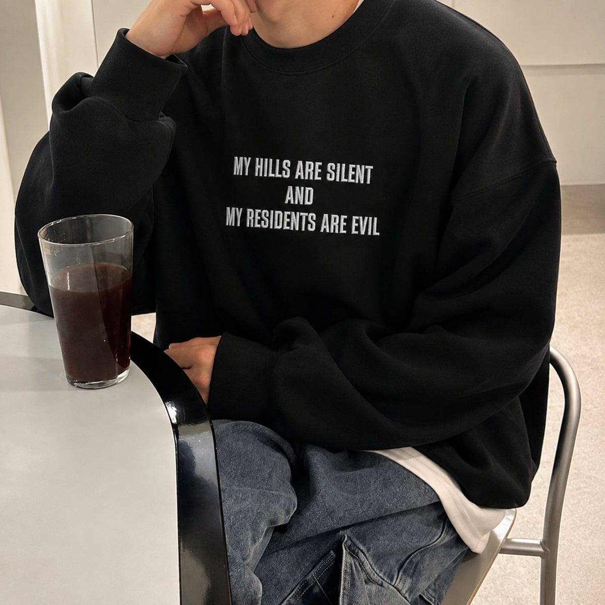 A person wearing a black oversized sweatshirt with white text that reads 'MY HILLS ARE SILENT AND MY RESIDENTS ARE EVIL', seated at a table with a glass of dark beverage.