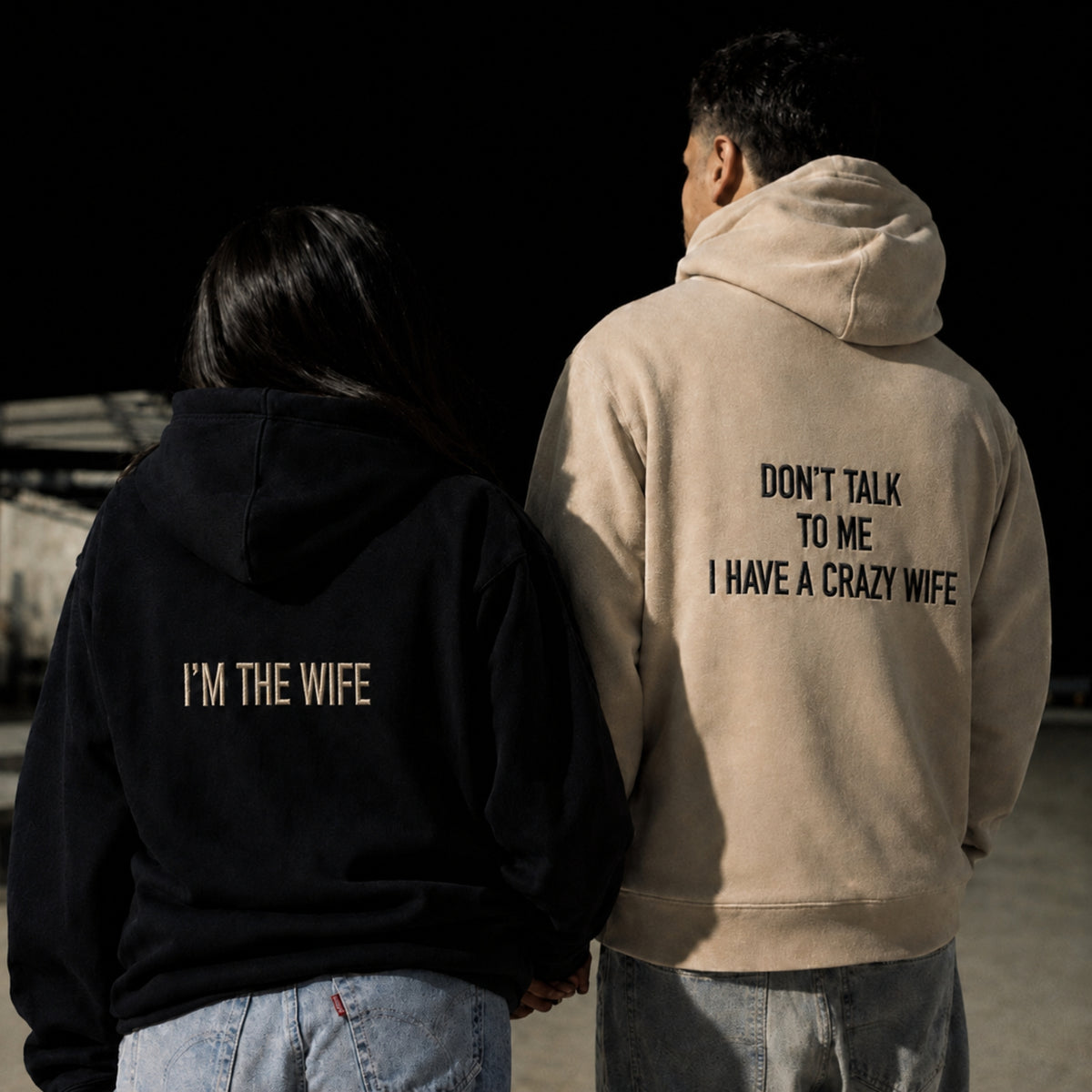 Two people standing side by side from behind, wearing matching graphic hoodies. The person on the left wears a black hoodie with 'I'M THE WIFE' printed in beige, and the person on the right wears a beige hoodie with 'DON'T TALK TO ME I HAVE A CRAZY WIFE' printed in black.