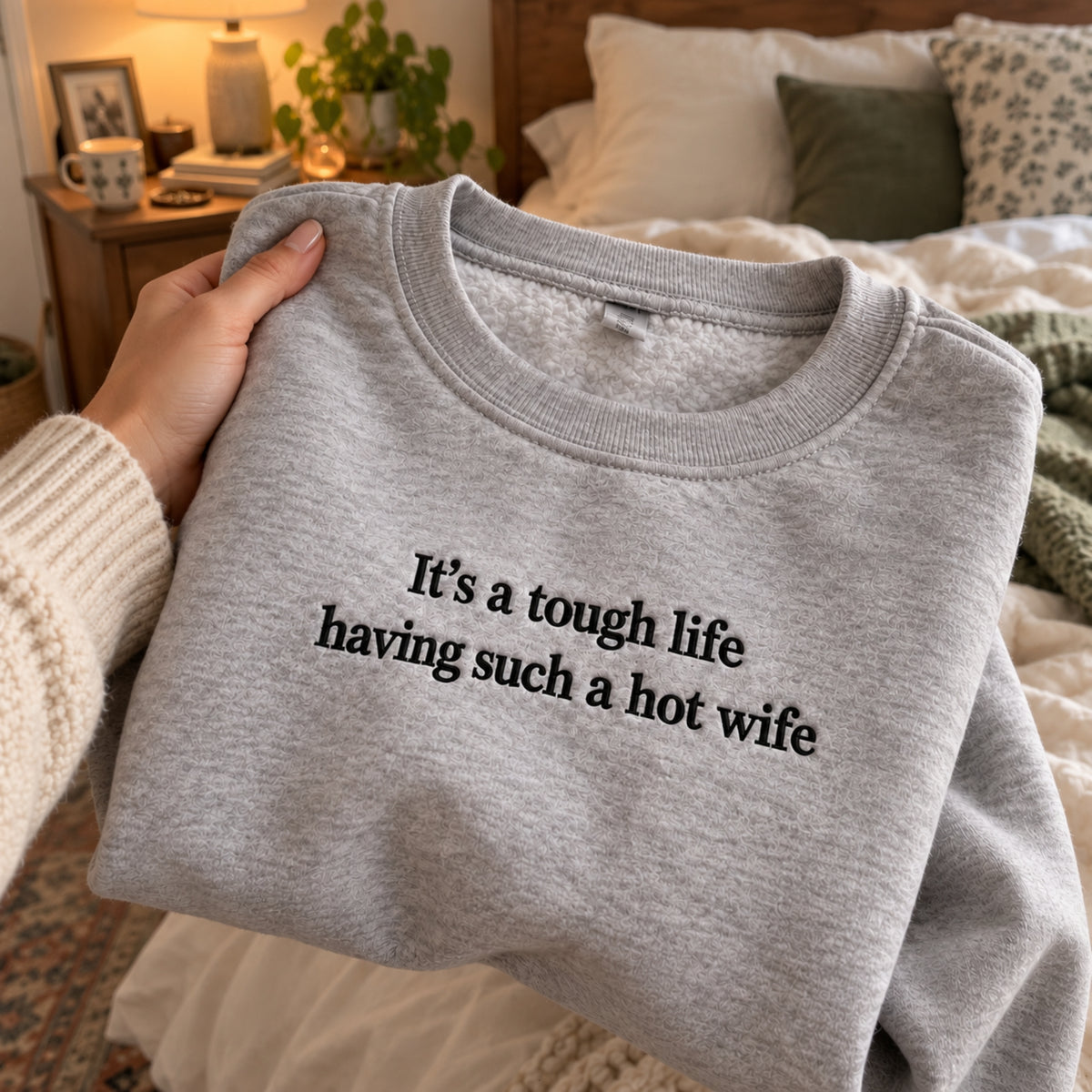 A folded heather gray sweatshirt with black text reading 'It's a tough life having such a hot wife' is being held by a person's hand in a cozy bedroom setting.