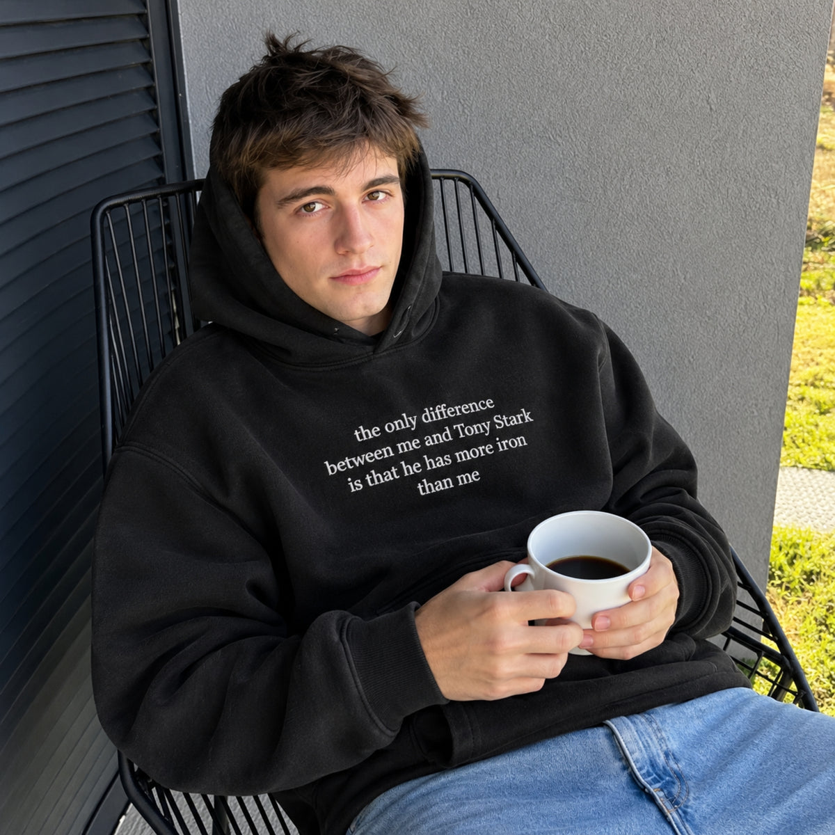 A man wearing a black hoodie with a white printed quote sits in a black wire chair, holding a white mug. The hoodie features the text: 'the only difference between me and Tony Stark is that he has more iron than me'.
