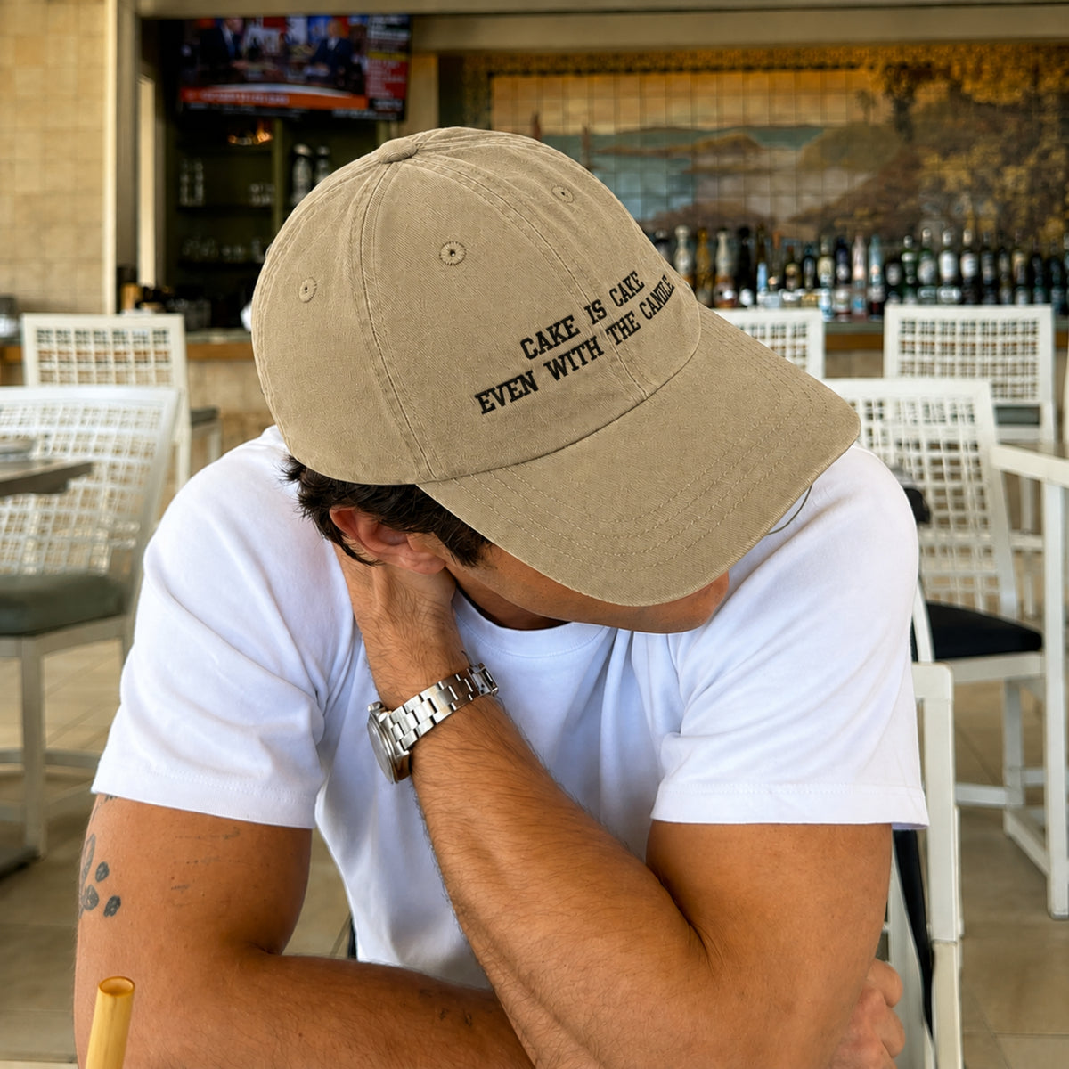 A man sitting at a cafe table wearing a beige baseball cap with the embroidered text 'CAKE IS GEEK EVEN WITH THE GEEK' and a white t-shirt.