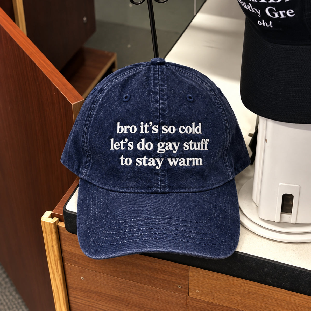 A navy blue washed baseball cap with white embroidered text reading 'bro it's so cold let's do gay stuff to stay warm', displayed on a wooden counter next to another black cap.
