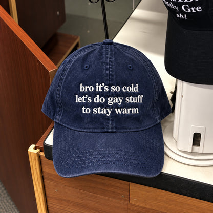 A navy blue washed baseball cap with white embroidered text reading 'bro it's so cold let's do gay stuff to stay warm', displayed on a wooden counter next to another black cap.