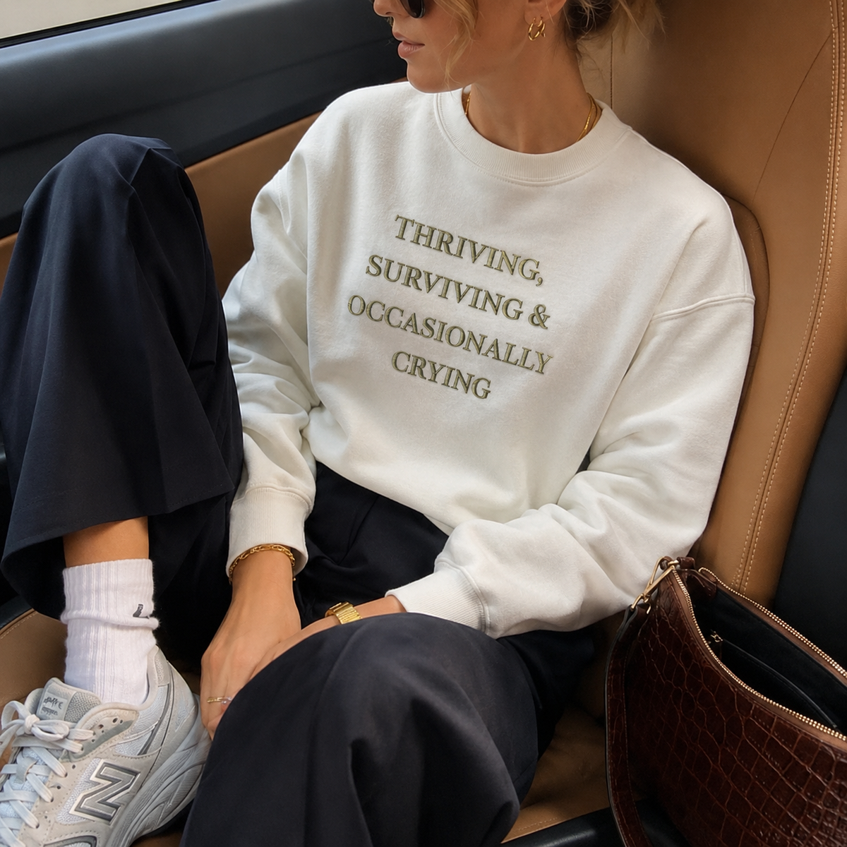 A woman sitting in a tan leather car seat wearing a white crewneck sweatshirt with the text 'THRIVING, SURVIVING & OCCASIONALLY CRYING' printed in olive green. She is also wearing dark navy pants, white socks, and New Balance sneakers.