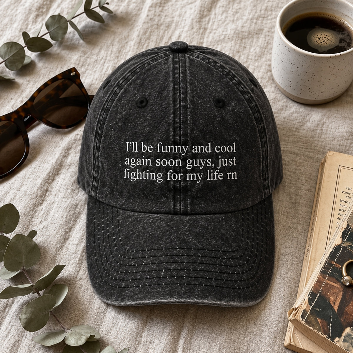 A dark washed baseball cap with white embroidered text reading 'I'll be funny and cool again soon guys, just fighting for my life rn', styled in a flat lay with sunglasses, a cup of coffee, eucalyptus leaves, and an open book on a light fabric background.