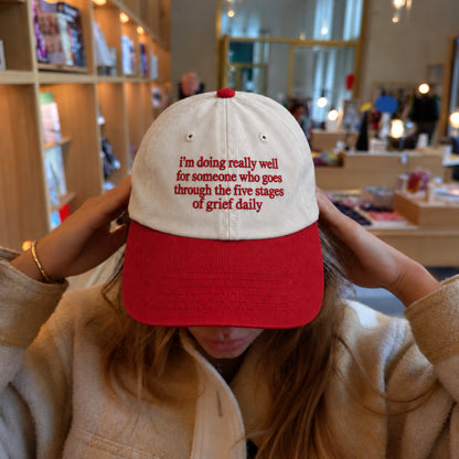 A person wearing a two-tone baseball cap with a red brim and red embroidered text on a cream crown, photographed in a retail store setting.