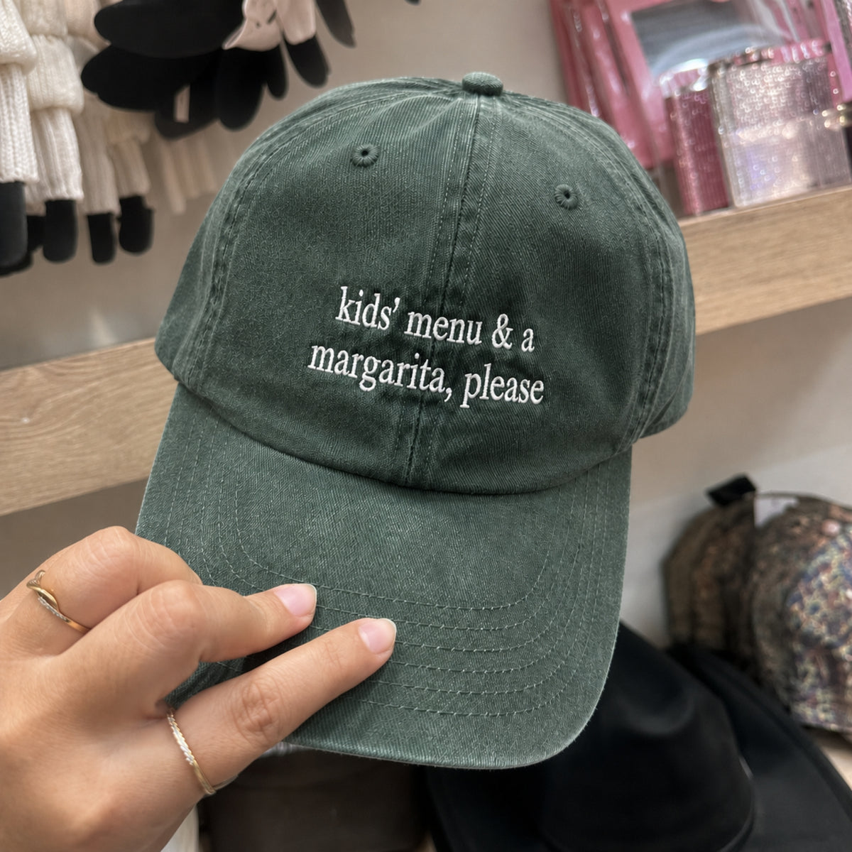 An olive green cotton baseball cap with white embroidered text reading 'kids' menu & a margarita, please' held by a hand in a retail store setting.