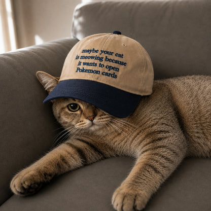 A brown tabby cat lies on a gray couch wearing a beige and navy baseball cap with embroidered text that reads, 'maybe your cat is meowing because it wants to open Pokemon cards.'.