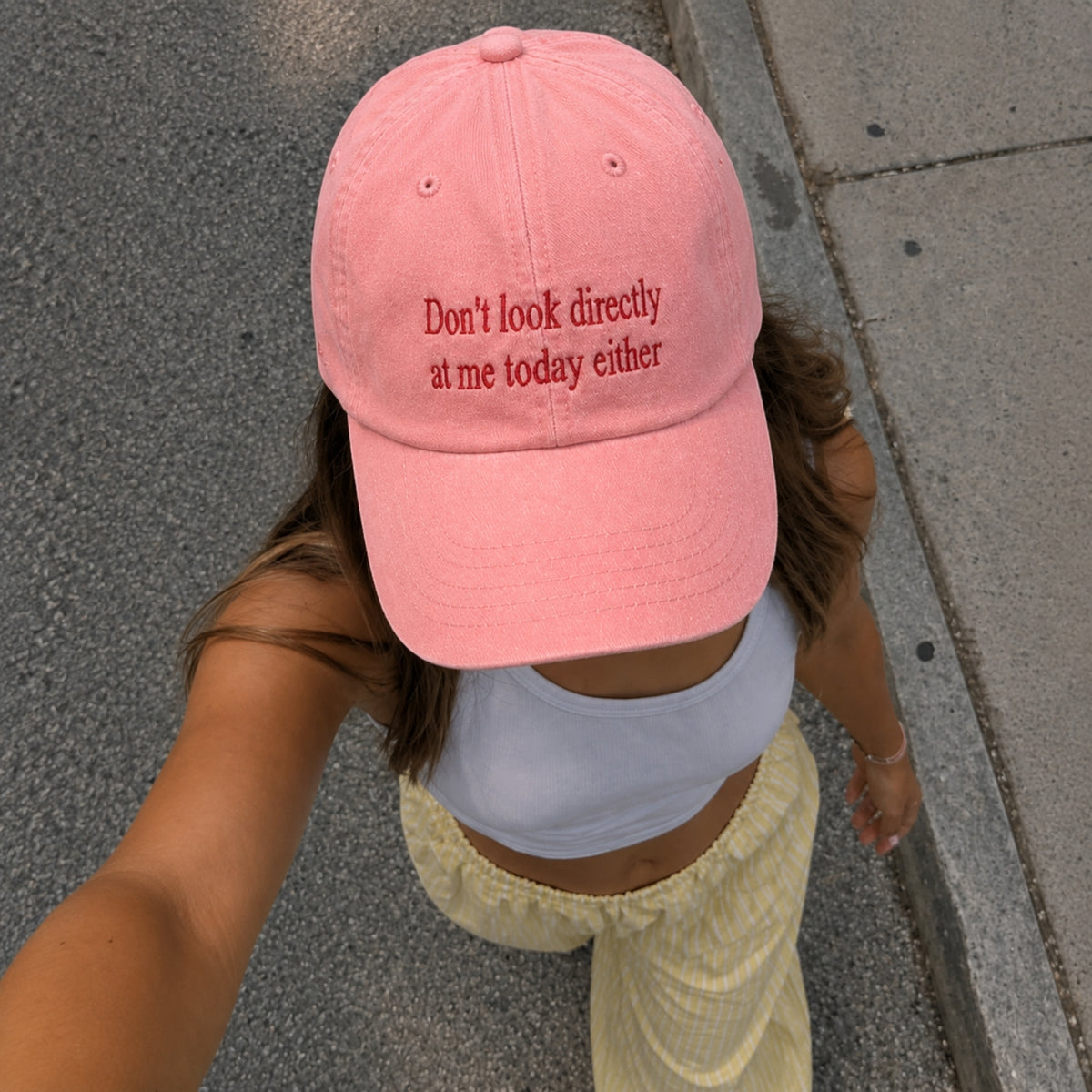 A person wearing a pink baseball cap with red embroidered text that reads 'Don't look directly at me today either' while walking outdoors on a paved street.